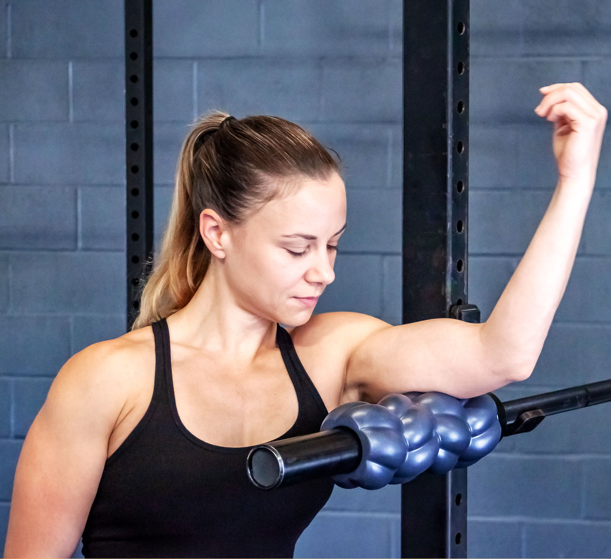 A fit woman rolling her triceps muscles with a barbell roller in a squat rack.